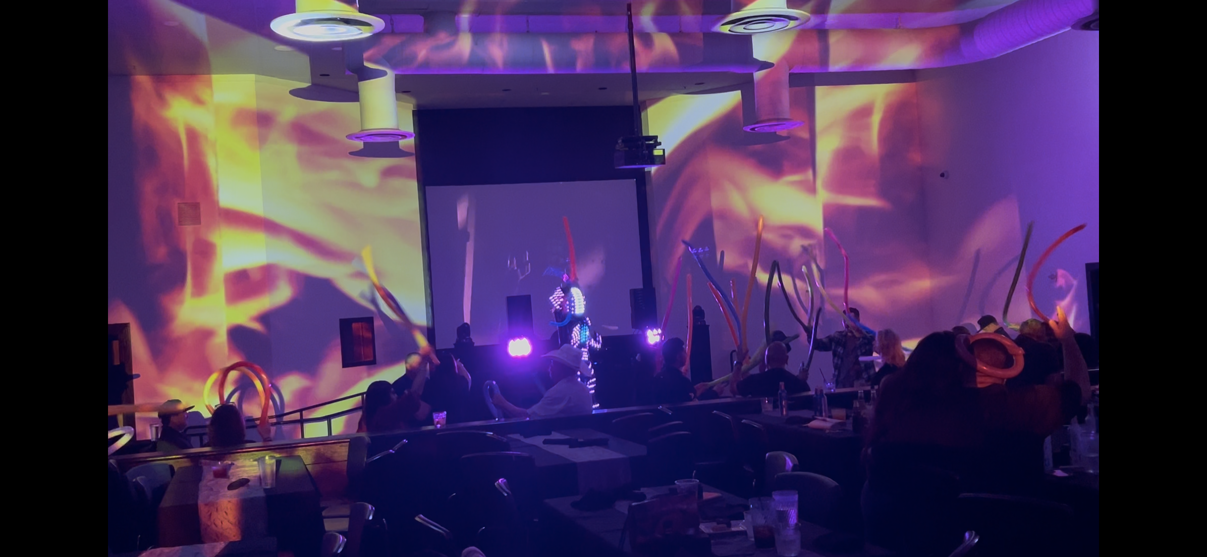 Fire-inspired projections cover the surrounding walls and overhead surfaces while an illuminated-costume performer entertains guests at a private event.